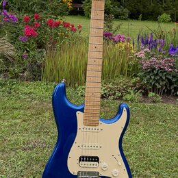 Modrá elektrická kytara typu Stratocaster s krčkem a hmatníkem ze světlého dřeva. Na hlavě je viditelné logo Fender. Kytara má krémový pickguard, tři…