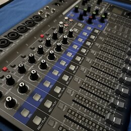 Šedý mixážní pult s modrými prvky a řadou posuvníků. Na panelu jsou viditelné nápisy jako GAIN, COMP, SEL, REC/PLAY a MUTE. Zařízení obsahuje početné…