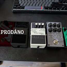Pět efektových pedálů v řadě na černém povrchu. Zleva je červený pedál, černý pedál s nápisem boss a bílý pedál s nápisy Noise Suppressor NS-2 a boss…