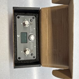 Černý elektronický přístroj v kartonové krabici. Na předním panelu jsou dva kovové konektory, tři stříbrná tlačítka a malý displej. Jsou zde nápisy U…