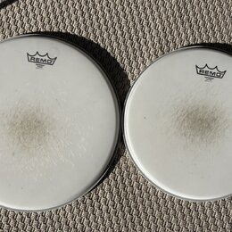 Dvě bílé bubnové kožehy značky REMO. Obě mají v horní části logo s nápisem REMO a pod ním text EMPEROR COATED MADE IN U.S.A. Povrchy kožehů vykazují…