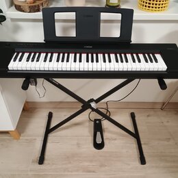 Černý digitální klavír značky YAMAHA umístěný na černém X-stojanu. Nástroj má bílé a černé klávesy a v horní části je připevněn černý notový pult s n…