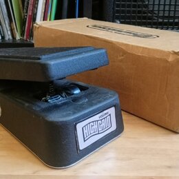 Černý volume pedál s nápisem VOLUME HIGH GAIN na bílém štítku. Vedle pedálu je umístěna hnědá kartonová krabice s nápisem GCB80 a čárovým kódem. Povr…