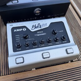 Stříbrný kytarový předzesilovač s nápisy AMP 1 a BluGuitar TONE INNOVATION. Na panelu jsou ovládací prvky s popisky VOLUME, GAIN, MASTER, BASS, MIDDL…