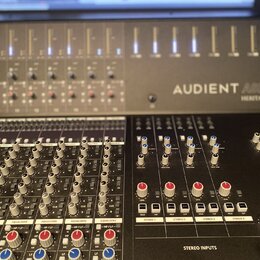 Fotografie zachycuje část míchacího pultu s nápisem AUDIENT ASP 4816 HERITAGE EDITION. Zařízení má šedý kovový povrch a je vybaveno řadou posuvníků,…