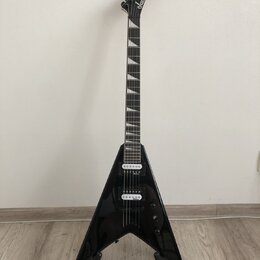 Černá elektrická kytara s tělem ve tvaru V umístěná na stojanu. Na hlavě hmatníku je bílý nápis Jackson. Kytara má dva humbuckery, jeden ovládací kno…