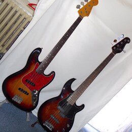 Dvě elektrické basové kytary s tmavě hnědým sunburst povrchem a hnedým hmatníkem. Levý nástroj má červený pickguard s texturou a na hlavě nápis Fende…