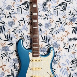 Modrá elektrická kytara s bílým perleťovým pickguardem a zlatými hardwaremi. Na hlavě hmatníku je nápis Squier STRATOCASTER. Hmatník je tmavého dřeva…