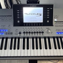 Stříbrný digitální klavír s nápisy YAMAHA a Tyros 5. Nástroj má v horní části displej, na kterém je zobrazen text Tyros 5 a další systémové informace…