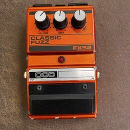 Oranžový efekt pedál s nápisy CLASSIC FUZZ a FX52. Na horní části jsou tři černé ovládací knoflíky s popisky VOLUME, TONE a FUZZ. V dolní části je lo…