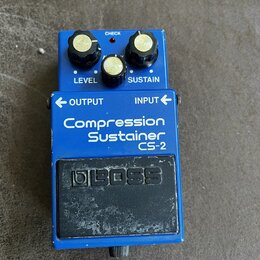 Modrý efekt pedál s nápisy LEVEL, SUSTAIN, OUTPUT a INPUT. Na přední straně je bílý text Compression Sustainer CS-2 a černé logo BOSS. Zařízení vykaz…
