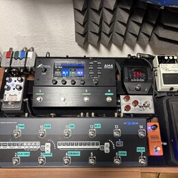 Sestava efektů na černém pedalboardu. V horní části jsou umístěny jednotky Fractal Audio AM4, Shure s displejem a bílý pedál Noise Suppressor NS-2. V…