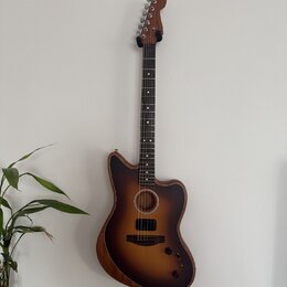 Akustická kytara s vyřezem v korpusu a tmavě hnědým přechodovým lakováním (sunburst). Na hlavě hmatníku je viditelné logo Fender. Nástroj visí na stě…