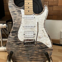 Elektrická kytara typu Stratocaster s tělem z vyvzorovaného dřeva v šedohnědé barvě a hmatníkem ze světlého javoru. Nástroj má bílý pickguard s perle…