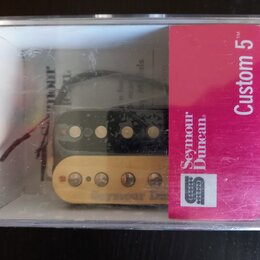 Průhledný plastový obal obsahuje dva elektrické snímače typu humbucker. Horní snímač je černý a dolní krémový, oba mají viditelné polosnímače. Na čer…