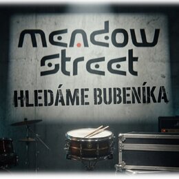 Fotografie zobrazuje buben s položenými paličkami, část činelu na stojanu a černý transportní case. V pozadí je zeď s velkým nápisem „meadow street“…