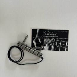 Kovový tremolo systém pro kytaru s černým přívodním kabelem. Vedle nástavce leží vizitka s nápisy BRENNER™, WWW.BRENNERUSA.COM, PRECISION GUITAR PROD…