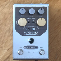 Stříbrný efekt pedál s nápisy MAGMA57 a Origin Effects. Horní část obsahuje tři ovládací knoflíky s popisky OUTPUT, TONE a DRIVE, dva zlaté knoflíky…