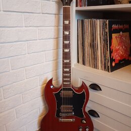 Tmavě červená elektrická kytara s černým pickguardem a hnedým hmatníkem. Na hlavě je nápis Gibson a pod ním označení SG. Tělo má charakteristický dvo…