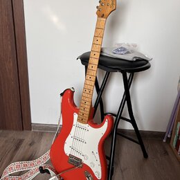 Červená elektrická kytara typu Stratocaster s bílým pickguardem a světle hnědým hmatníkem. Na hlavě kytary je nápis Squier by Fender STRATOCASTER. Ná…