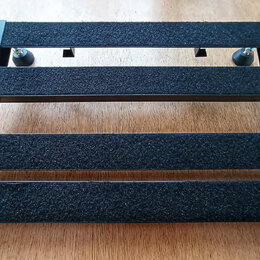 Černý obdélníkový pedalboard s třemi vodorovnými lištami potaženými černým hrubým povrchem. Boční rám je vyroben z černého materiálu s viditelnými ot…
