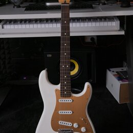 Bílá elektrická kytara typu Stratocaster s hnědým pickguardem a hnedým hmatníkem. Na hlavě je vidět nápis Fender. Kytara má tři snímače, tři ovládací…