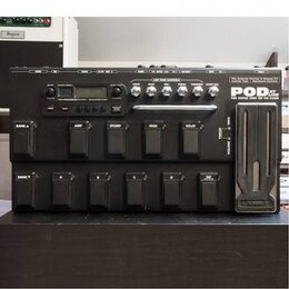 Černý kytarový procesor Line 6 POD XT Live. Na horním panelu jsou ovládací knofíky, displej a nápisy AMP TONE CONTROLS, DRIVE, BASS, MID, TREBLE, PRE…