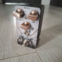 Efektový pedál s černým šasi a stříbrno-šedou přední deskou. Na desce je nápis TONE SLAVE a logo VB Pedals, doplněné motivem postavy v řetězech. Pedá…
