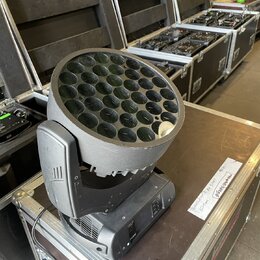 Černý LED reflektor typu moving head s kulatou hlavou obsahující mřížku s mnoha malými čočkami. Zařízení stojí na hliníkovém transportním kufru v pro…