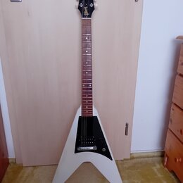 Bílá elektrická kytara s tělem ve tvaru V a hnědým hmatníkem. Na hlavě je viditelné zlaté logo Gibson. Tělo má černý plech s dvěma snímači a ovládací…