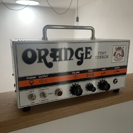 Kytarový zesilovač značky Orange, model Tiny Terror, v bílém provedení s kovovou rukojetí. Na předním panelu je velký černý nápis ORANGE a menší text…