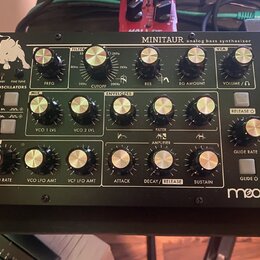 Černý analogový basový syntetizátor s nápisem MINITAUR a analog bass synthesizer. V levém dolním rohu je bílé logo moog. Přístroj obsahuje několik řa…