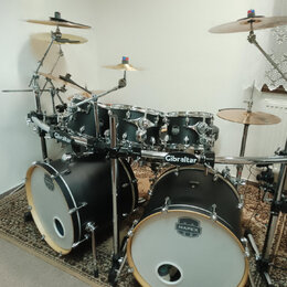 Černá bicí souprava s bílými لديmi a chromovaným kováním. Na basových bubnech je logo MAPEX, na tomarech jsou vidět loga Mapex a na nosném systému ná…
