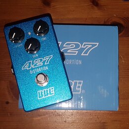 Modrý efekt pedál s glitrovým povrchem a tři ovládací knoflíky. Na pedálu jsou bílé nápisy 427 DISTORTION, LEVEL, GAIN, TONE a logo BBE. Vedle pedálu…