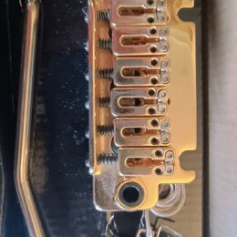 Zlatý Tremolo bridge pro elektrickou kytaru s šestnácti sedly a součástí je páka. Kovový povrch vykazuje známky opotřebení, drobné škrábance a oxidac…