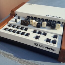 Bílý elektronický klávesový syntetizátor s dřevěnými bočními panely. Na horním panelu je nápis OB-3 a logo Oberheim, v dolní části je opět logo Oberh…