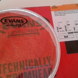 Průhledná plastová bubna v bílém obruči položená na červeném kartonovém obalu. Na bubně je černý nápis EVANS G2 LEVEL 360 A D'ADDARIO INNOVATION. Na…