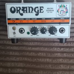 Bílý kytarový zesilovač s kovovou rukojetí a černými nohami. Na horním panelu je nápis ORANGE, MICRO TERROR a 20W RMS spolu s barevným logem. Ovládac…