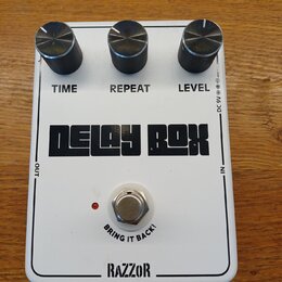 Bílý efekt pedál s nápisem DELAY BOX a značkou RAZZOR v dolní části. Na horní straně jsou tři černé ovládací knoflíky s popisky TIME, REPEAT a LEVEL.…