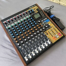 Míchací pult Model 12 značky TASCAM v černém provedení s dřevěnými bočními panely. Zařízení obsahuje více vstupních kanálů s řadou potenciometrů, pře…