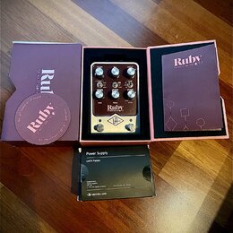Kytarový pedál v barvě stříbrné a černé, umístěný v originální fialové krabici. Na pedálu je nápis Ruby, TOP BOOST AMPLIFIER '63 a logo UA v kosočtve…