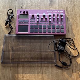 Fialový sampler s nápisy electribe sampler a KORG. Zařízení obsahuje displej, několik otočných knoflíků a mřížku s čtvercovými padovými tlačítky. K p…