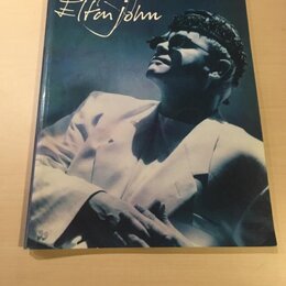 Fotografie znázorňuje obal CD nebo album s portrétem muže v brýlích a bílém saku. V horní části je nápis „the very best of Elton John“. Předmět leží…