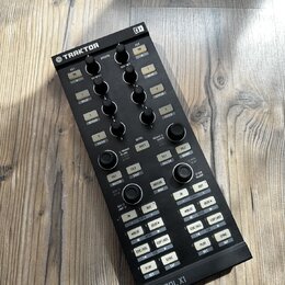 Černý DJ kontrolér s nápisy TRAKTOR a KONTROL X1. Zařízení obsahuje několik řad knofíků, posuvníků a bílých tlačítek s popisky jako DELAY, REVERB, FI…