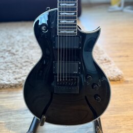 Černá elektrická kytara s lesklým povrchem a bílým lemováním korpusu. Na snímači je viditelný nápis EMG. Kytara má dva humbuckery, tři ovládací knofí…