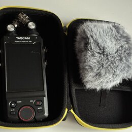Černý digitální záznamový zařízení v žlutém přepravním pouzdře. Na horní části přístroje je bílý nápis TASCAM a pod ním Portacapture X8. V pravé část…