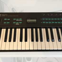 Černý digitální klavír s bílými a černými klávesami. Na horním panelu je nápis YAMAHA DX21, malý displej a řada zelených tlačítek. Nástroj vykazuje m…
