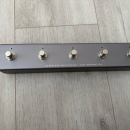 Šedý MIDI footswitch v kovovém obalu s pěti stříbrnými spínači. Na spodní hraně je bílý nápis DARKGLASS ELECTRONICS - MIDI FOOTSWITCH. Povrch zařízen…