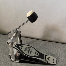 Stříbrný basový pedál s černým šlapadlem. Na šlapadle jsou nápisy IRON COBRA 200 a logo TAMA. Povrch vykazuje mírné známky používání v podobě drobnýc…