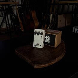 Bílý efekt pedál s nápisem FUZZ a logem JHS SERIES. Na horní straně jsou tři ovládací knoflíky s popisky VOLUME, BASS a FUZZ. Vedle pedálu je umístěn…